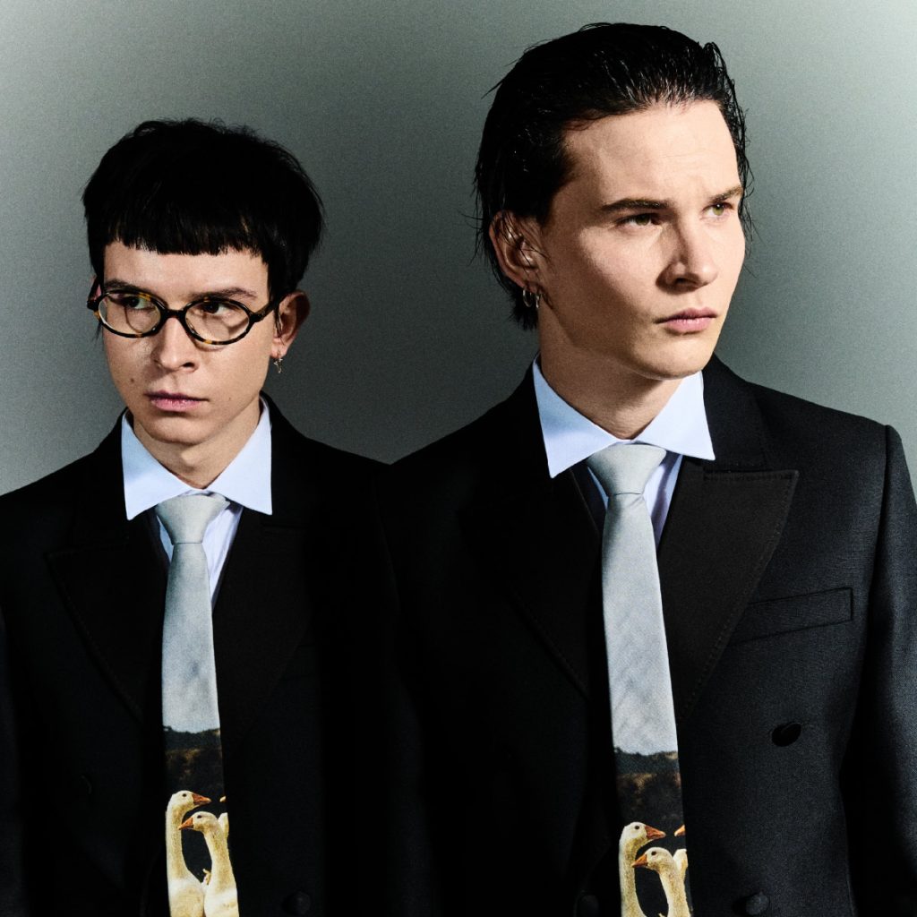 Zdjęcie przedstawia dwóch młodych mężczyzn stojących obok siebie na neutralnym, szarym tle. Obaj mają na sobie czarne garnitury, białe koszule oraz jasne krawaty z nadrukiem przedstawiającym dwa ptaki. Mężczyzna po lewej ma krótkie, ciemne włosy i okrągłe okulary, patrzy lekko w bok. Mężczyzna po prawej ma zaczesane do tyłu, ciemne włosy i spogląda w dal z poważnym wyrazem twarzy. Całość ma elegancki, minimalistyczny charakter.