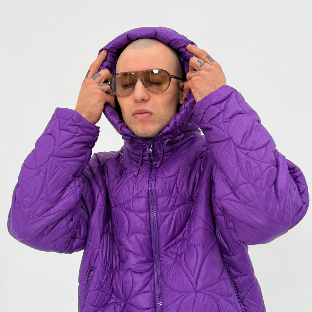 Mężczyzna w fioletowej pikowanej kurtce z kapturem i okularach przeciwsłonecznych, trzyma dłonie przy kapturze; minimalistyczne, jasne tło
