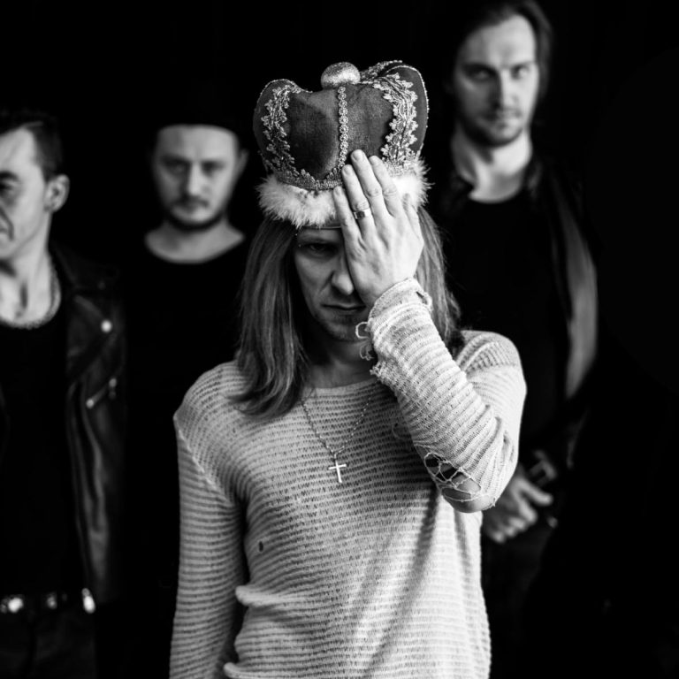 Czarno-biała fotografia przedstawiająca czterech mężczyzn stojących na ciemnym tle. Na pierwszym planie znajduje się mężczyzna w koronie obszytej futrem, który zasłania dłonią połowę twarzy. Ma długie włosy, na szyi wisi krzyżyk, a ubrany jest w jasną, prążkowaną koszulkę z długim rękawem. Za nim stoją trzej inni mężczyźni w ciemnych ubraniach, lekko rozmyci, patrzący w stronę obiektywu. Całość ma surowy, nastrojowy charakter.